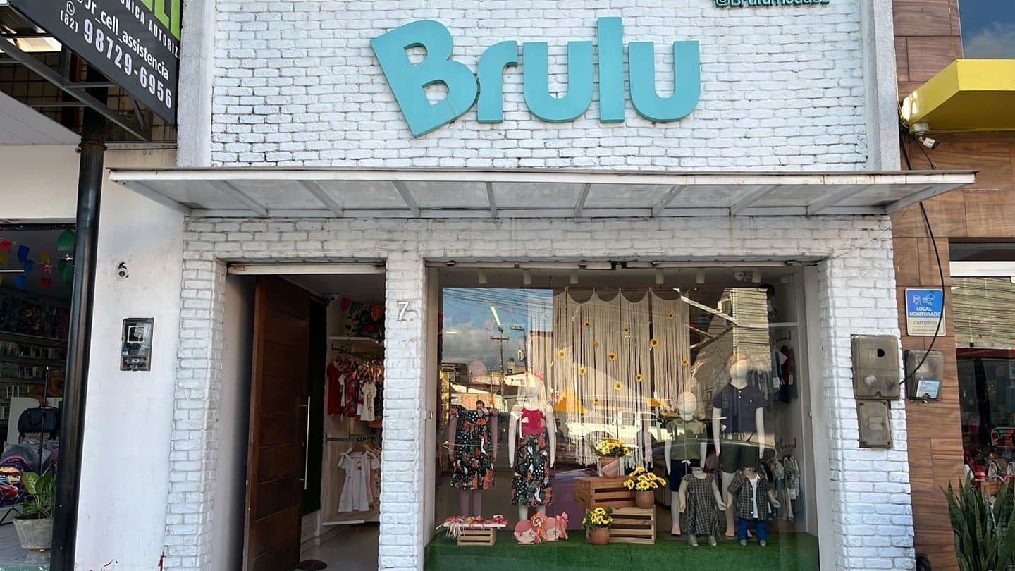 Fachada da loja Brulu: parede de tijolo branco com o letreiro turquesa Brulu e a vitrine com manequins infantis vestidos