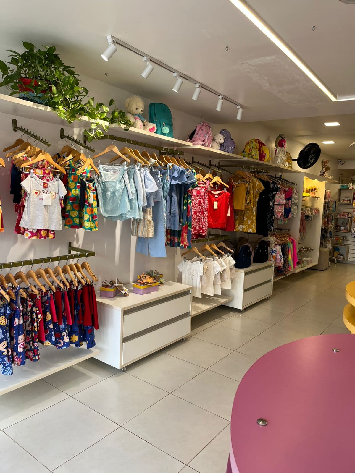 Araras da loja Brulu com vestidos, camisetas e shorts infantis organizados por cor, e mochilas nas prateleiras de cima
