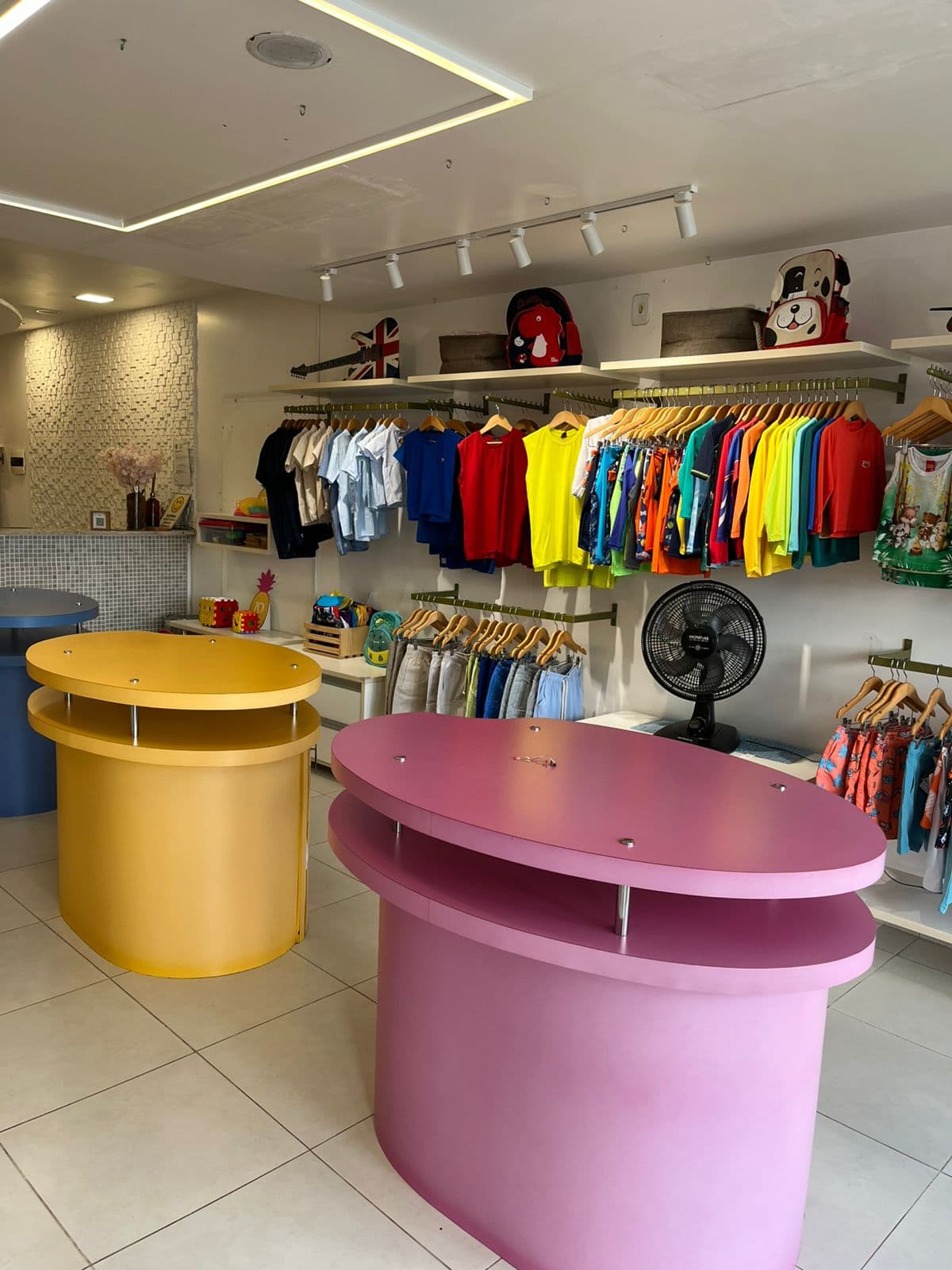Expositores redondos amarelo e rosa no meio da loja, com camisetas coloridas e bermudas juvenis ao fundo